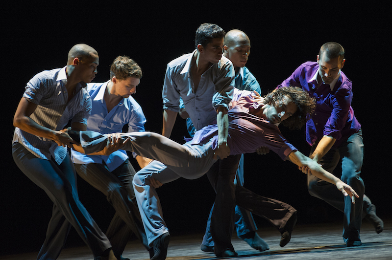 A group of male dancers huddle together to support another male dancer as he extends to the ground